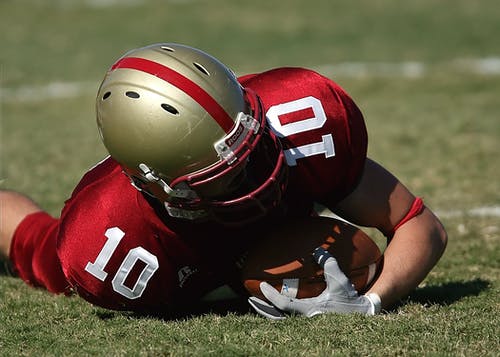 红色白色橄榄球球衣和灰色红色头盔举行足球和躺在绿色的草地上的男人 · 免费素材图片