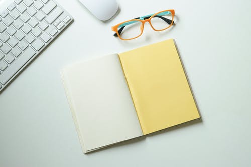 白色和黄色笔记本，放在键盘和眼镜附近 · 免费素材图片