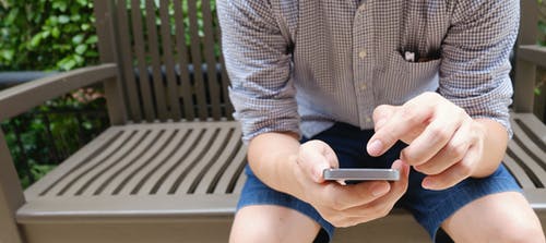 男人穿着棕色和白色格子运动衬衫坐在棕色的长椅上，白天使用智能手机 · 免费素材图片
