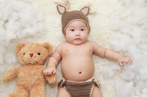 婴儿躺在棕熊毛绒玩具旁边的白色垫子上 · 免费素材图片