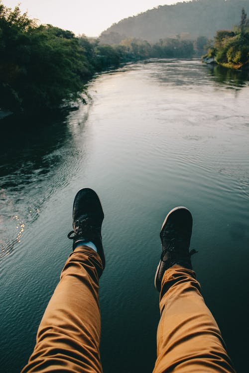 黑色运动鞋和棕色的裤子，坐在河边的人 · 免费素材图片