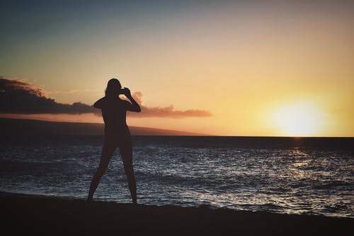 在黄金时段在海边附近拿着相机的女人的剪影 · 免费素材图片