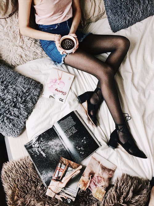 一个女人躺在床上被毛皮枕头和杂志包围的同时拿着白色杯子与黑色液体的平板摄影 · 免费素材图片