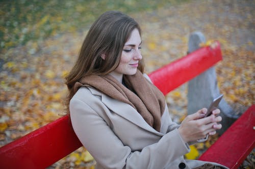 米色外套拿着智能手机，坐在长椅上的女人 · 免费素材图片