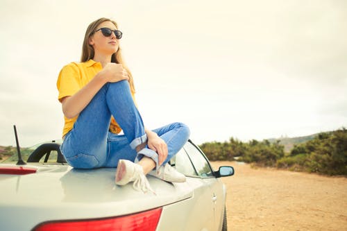 女人穿着黄色的衬衫和蓝色的牛仔牛仔裤坐在银车上 · 免费素材图片