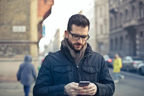 男人穿着黑色拉链夹克拿着智能手机包围灰色混凝土建筑 · 免费素材图片