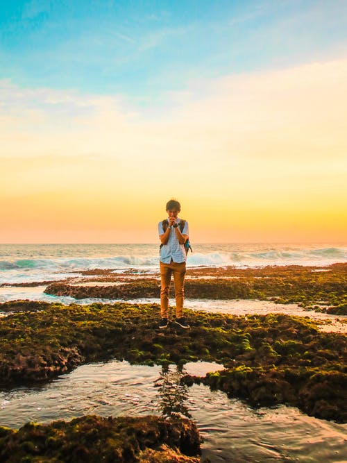 在黄金时段，男人站在海滩附近的岩石上 · 免费素材图片