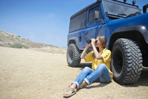 使用双筒望远镜时坐在吉普车旁边的土壤上的女人 · 免费素材图片