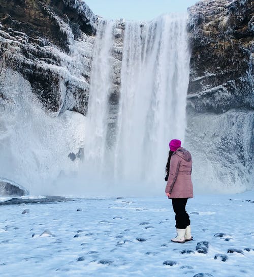 女人穿着粉红色的雪外套站在冰冷的瀑布前充满了雪的字段上 · 免费素材图片