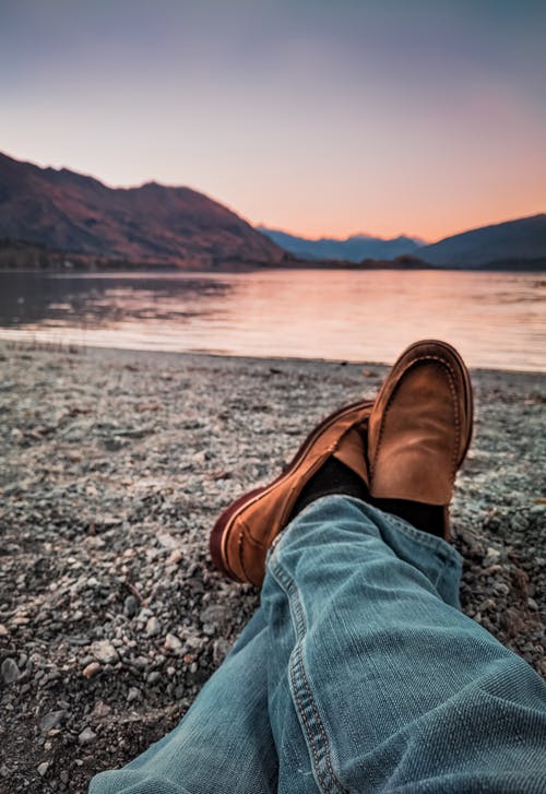 坐在灰色的沙子旁边的穿着蓝色牛仔裤和棕色皮革便鞋的人 · 免费素材图片
