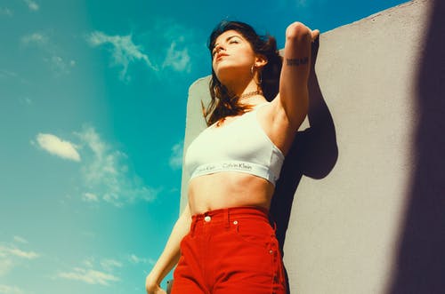 白色的卡尔文·克莱因运动胸罩和红色底部倾斜在灰色的天空下的灰色墙上的女人 · 免费素材图片