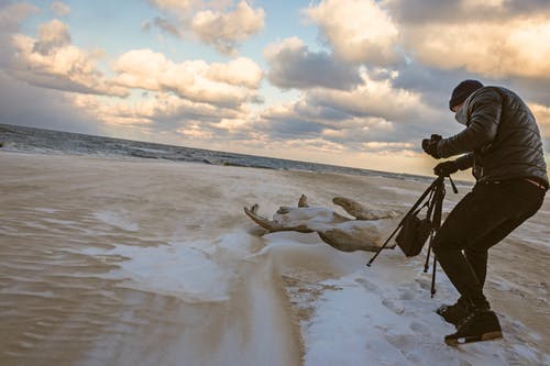 手持黑色相机三脚架在海边散步的黑色泡泡夹克的男人 · 免费素材图片