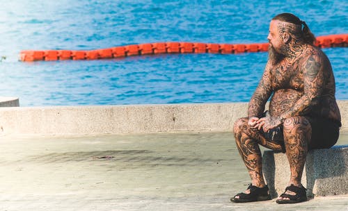 坐在水附近的灰色混凝土地板上的纹身的男人 · 免费素材图片