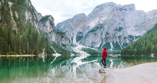 身穿红色连帽衫的男子站在水体附近与山景 · 免费素材图片