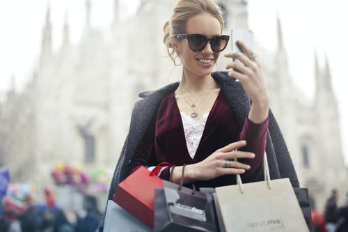 栗色的长袖上衣，白天拿着智能手机与购物袋的女人 · 免费素材图片