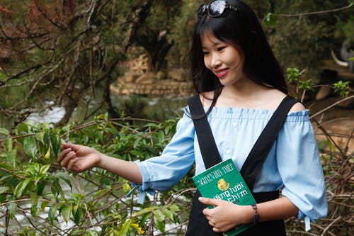 女人穿着蓝色露肩上衣拿着本书旁边绿叶植物 · 免费素材图片