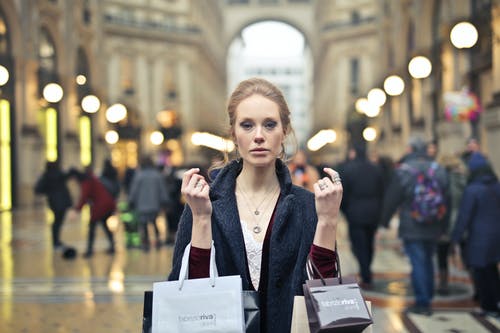 女人穿着黑色大衣，拿着各种颜色的购物袋，在建筑物上 · 免费素材图片