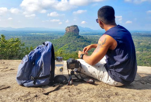 坐在背包，水壶和相机旁边的灰色悬崖山上的人 · 免费素材图片