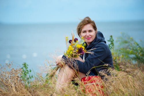 女人坐在草捧着鲜花，穿着黑色连帽衫 · 免费素材图片