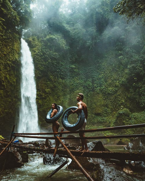 两人背着黑色充气游泳池浮在瀑布附近的棕色木桥上 · 免费素材图片