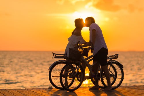 日落期间在河码头上拿着自行车时亲吻女人的男人穿着白色t恤的摄影 · 免费素材图片