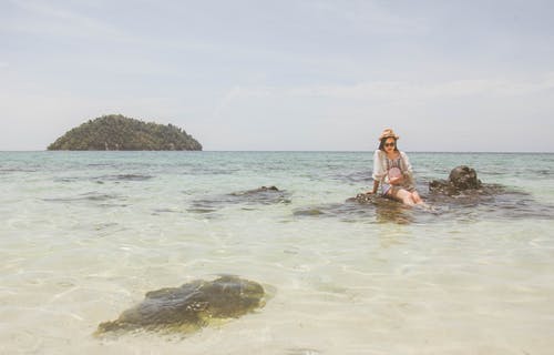 女人穿着白色上衣，坐在水体的棕色岩石上 · 免费素材图片
