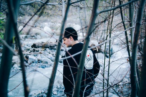 穿黑色运动衫站在河和光秃秃的树木旁边的人 · 免费素材图片