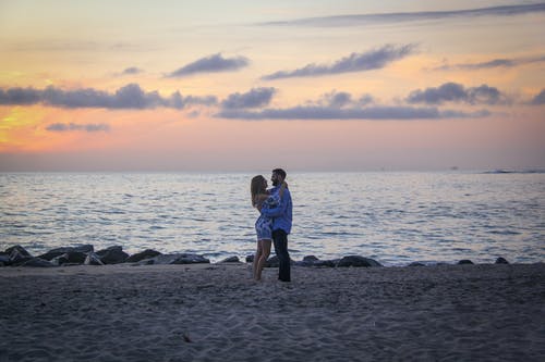 黄金时段，情侣在平静的水面附近的海滨站立 · 免费素材图片
