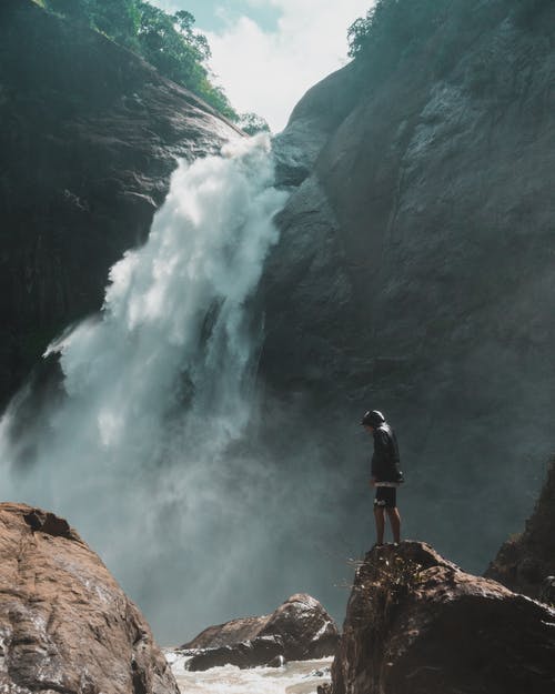 男子站在瀑布摄影前的棕色岩石悬崖上 · 免费素材图片