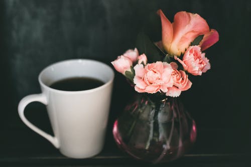 粉红色的康乃馨花和粉红色的玫瑰花朵，在杯咖啡旁边的透明玻璃花瓶里 · 免费素材图片
