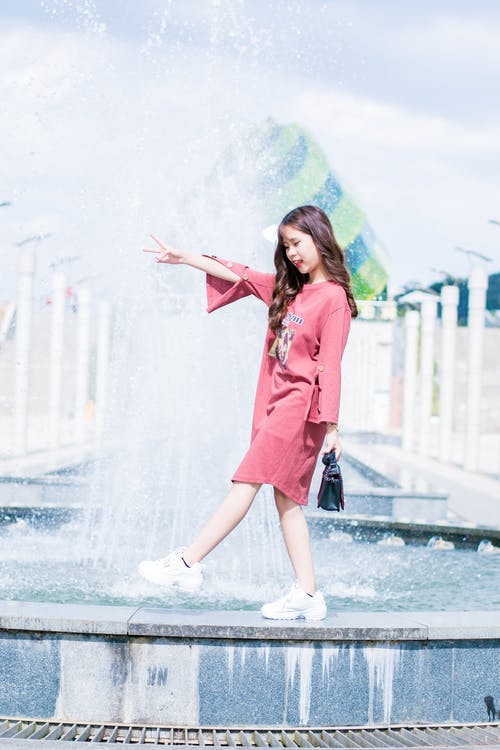 戴粉红色的长袖连衣裙，在白天散步在喷泉上的女人 · 免费素材图片