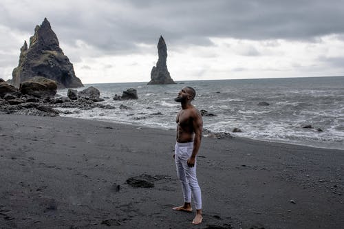 穿着白色牛仔裤的男人站在灰色的沙滩，在灰色的云彩下的灰色沙滩上 · 免费素材图片