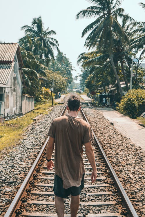 棕色衬衫的男人站在椰子棕榈树附近的火车轨道上 · 免费素材图片