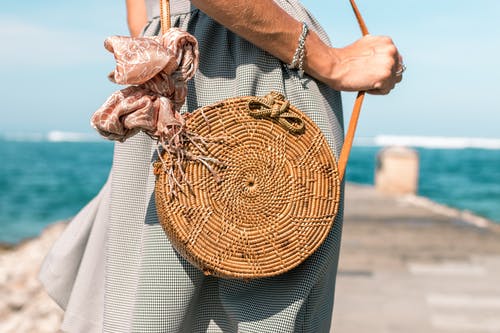 女人穿着灰色的裙子和圆形的棕色藤制斜挎包袋在木码头附近的水体 · 免费素材图片