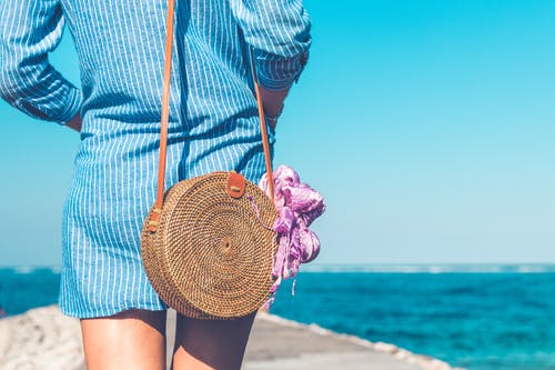 女人穿着蓝色和白色的条纹的裙子与棕色藤藤斜挎包袋附近海洋 · 免费素材图片
