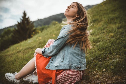 女人穿着蓝色牛仔夹克，坐在白天在树下蓝蓝的天空下的绿草附近 · 免费素材图片