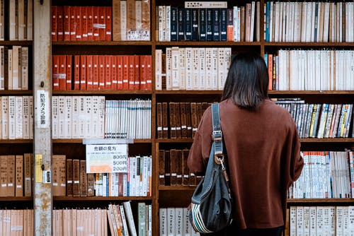 女人穿着棕色衬衫，黑色的皮包，在图书馆的书前 · 免费素材图片