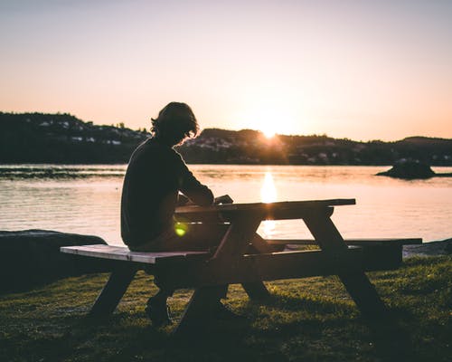 在日落期间坐在水边附近的野餐长凳上的黑色上衣的人的剪影 · 免费素材图片