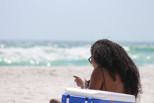 女人坐在岸边的蓝色和白色冷却箱旁边的沙滩上 · 免费素材图片