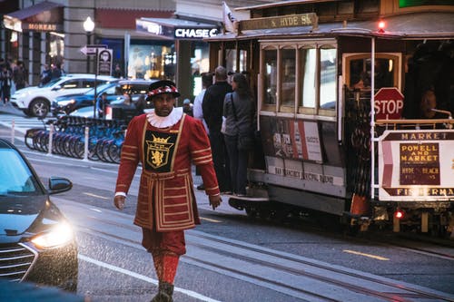 男人穿着红色和黄色的衣服，在白色和红色的电缆火车附近的混凝土路面上行走 · 免费素材图片