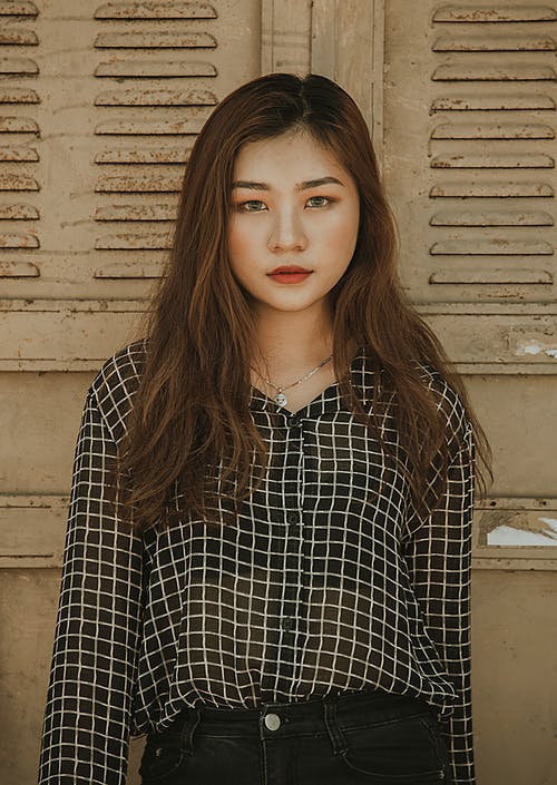 女人穿着白色和黑色的tattersall领口的纽扣衬衫站在棕色的墙附近 · 免费素材图片