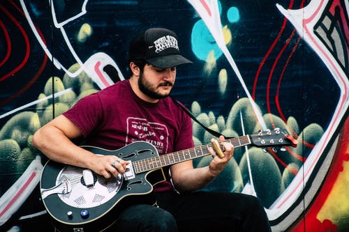穿红衬衫的男人，在黑色和绿色绘画壁附近玩谐振器吉他 · 免费素材图片