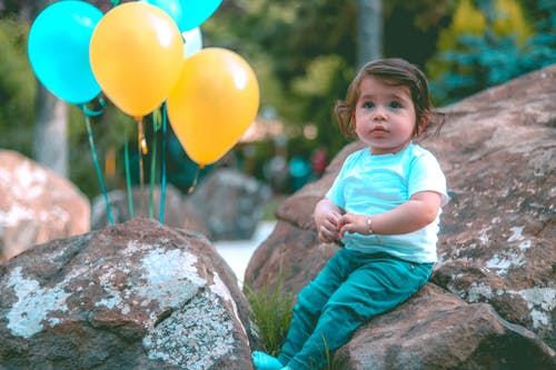 蹒跚学步的白衬衫，坐在黄色和蓝色的气球旁边的岩石上 · 免费素材图片
