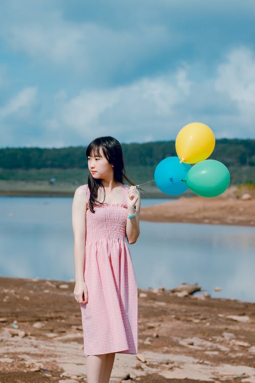 女人穿着粉红色的连衣裙，拿着三个气球在水体附近 · 免费素材图片