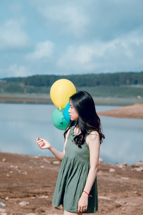 穿着绿色圆领无袖迷你连衣裙的女人拿着气球靠近水体 · 免费素材图片