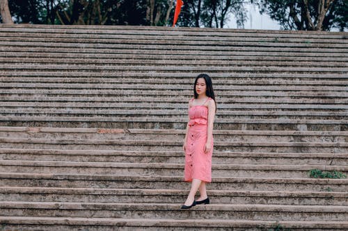 女人穿着粉红色的意大利面条皮带礼服站在灰色的混凝土楼梯上 · 免费素材图片