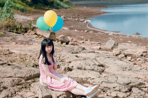 粉红色的厚皮带礼服，坐在岩石地形上拿着黄色和蓝色的气球的女人 · 免费素材图片