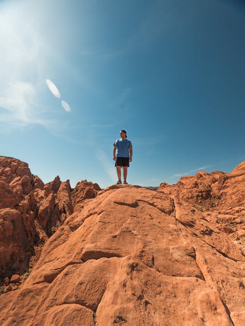 穿着蓝色衬衫和黑色短裤的男人站在晴朗的天空下的棕色岩层上 · 免费素材图片