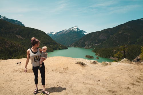 在山间遥远的湖中穿灰色背心抱婴儿的女人 · 免费素材图片