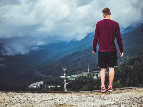 男人穿着栗色毛衣和黑色短裤站在山前 · 免费素材图片